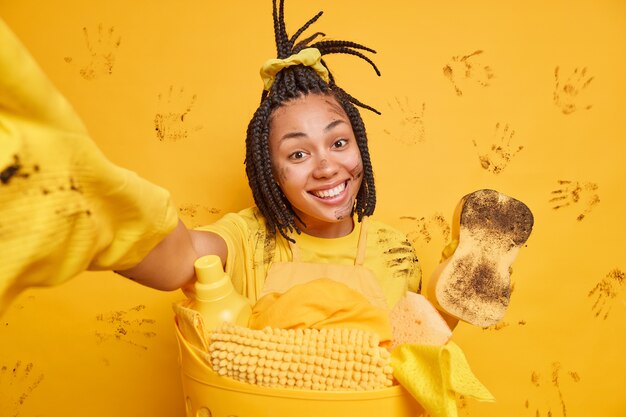 Une fille afro-américaine positive avec un sourire à pleines dents le visage sale s'étire le bras fait une photo d'elle-même en faisant la lessive à la maison essuie la saleté a le nettoyage de printemps pendant le week-end isolé sur un mur jaune