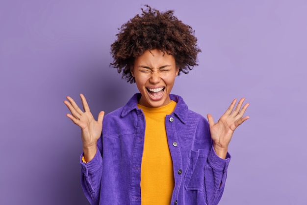 Une fille afro-américaine ravie s'exclame fort, garde les mains levées, se sent excitée et très heureuse d'entendre des nouvelles impressionnantes vêtue d'une veste en velours.