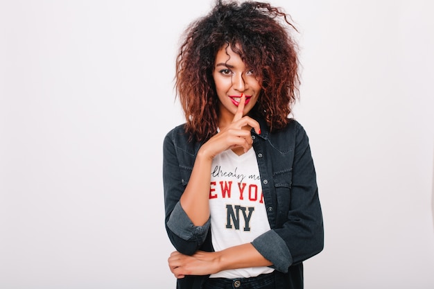 Fille afro-américaine sensuelle avec une coiffure frisée tendance portant une veste en jean et une chemise avec impression. Portrait intérieur d'un modèle féminin noir fascinant posant de manière ludique.