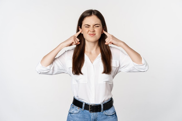 Une fille agacée ferme les oreilles à cause d'un son fort, inconfort d'entendre quelque chose de bruyant, debout gênée sur fond blanc.