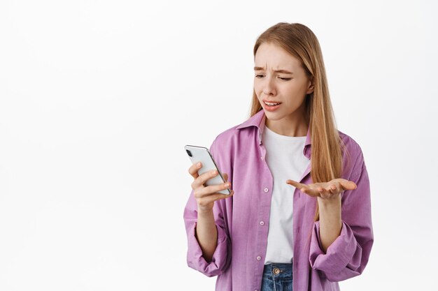 La fille a l'air confuse au smartphone, reçoit un message étrange au téléphone, se plaint, a l'air perplexe, debout sur fond blanc