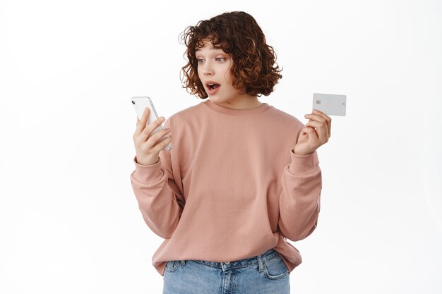La fille a l'air surprise de l'écran du téléphone portable lors de ses achats en ligne, tenant une carte de crédit, payant pour la livraison des commandes sur Internet, spot bon prix dans la boutique, debout sur blanc