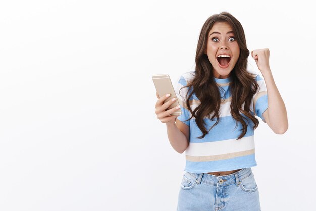 Une fille amusée reçoit des nouvelles fantastiques, une pompe de poing joyeusement, tient un smartphone souriant largement en criant hourra