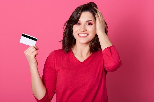 Fille avec une apparence agréable tenant une carte de crédit dans ses mains, gagne de l'argent à la loterie, une femme en tenue rouge a l'air heureuse, paie pour les achats par carte, en gardant la paume sur le troupeau.