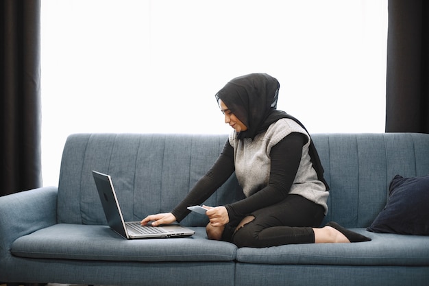 Fille arabe moderne en hijab utilisant un ordinateur portable à la maison, travaillant à distance assis sur un canapé dans le salon