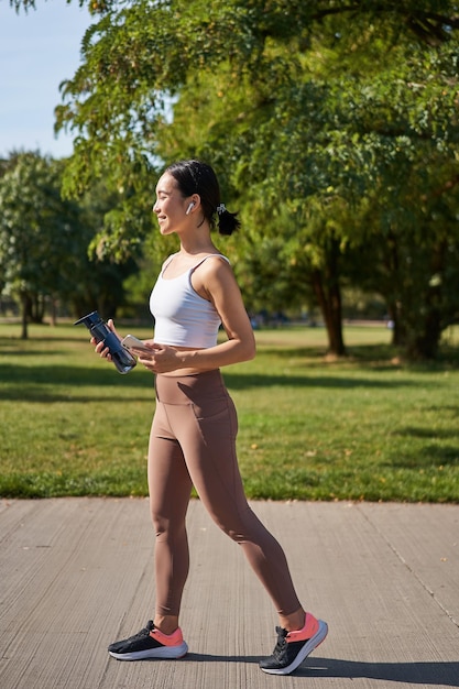 Fille asiatique active dans l'entraînement de vêtements de fitness dans le parc marchant dans des vêtements de sport avec smartphone et eau