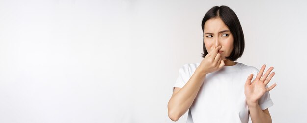 Une fille asiatique a l'air dégoûtée de rejeter le produit avec une mauvaise odeur ferme le nez de l'aversion et grince des dents debout sur fond blanc