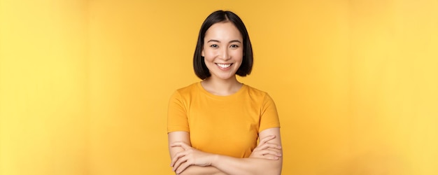 Une fille asiatique confiante croise les bras sur la poitrine en souriant et en s'affirmant debout sur fond jaune