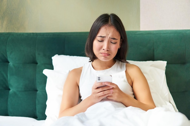 Fille asiatique déçue pleurant et pleurnichant se sentant bouleversée allongée dans son lit sur un oreiller tenant un téléphone portable