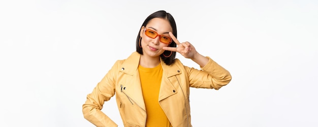 Fille asiatique élégante dans des lunettes de soleil a l'air cool et à la mode montre le geste de vsign de paix se dresse sur blanc b