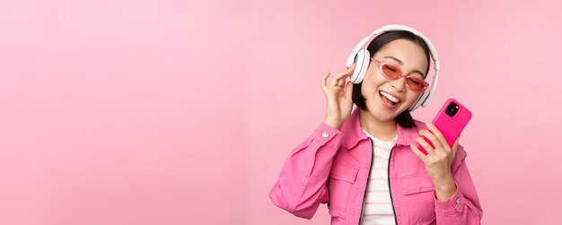 Fille asiatique élégante dansant avec un smartphone écoutant de la musique dans des écouteurs sur une application de téléphone mobile souriant et riant posant sur fond rose