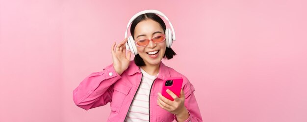 Fille asiatique élégante dansant avec un smartphone écoutant de la musique dans des écouteurs sur une application de téléphone mobile souriant et riant posant sur fond rose