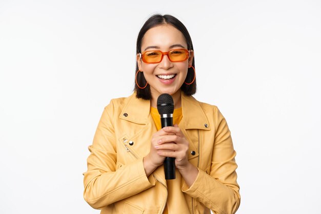 Fille asiatique élégante à lunettes de soleil chantant des chansons avec microphone tenant un micro et dansant au karaoké posant sur fond blanc