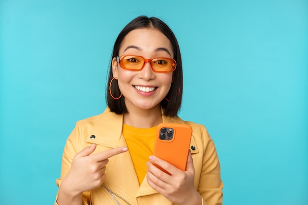 Fille asiatique enthousiaste dans des lunettes de soleil élégantes pointant le doigt sur un smartphone montrant un téléphone portable debout sur fond bleu