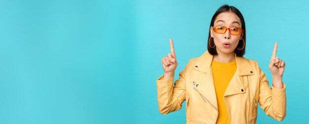 Une fille asiatique enthousiaste dans des lunettes de soleil pointe les doigts vers le haut montre une bannière ou un logo sur le dessus se dresse sur fond bleu Copier l'espace