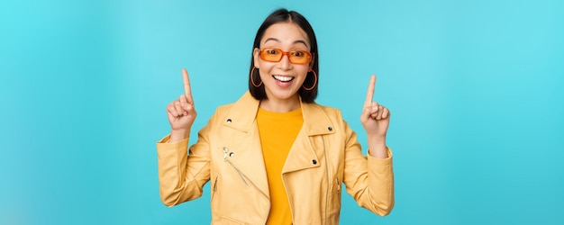 Une fille asiatique enthousiaste en lunettes de soleil pointe les doigts vers le haut montre une bannière ou un logo sur le dessus se dresse sur fond bleu
