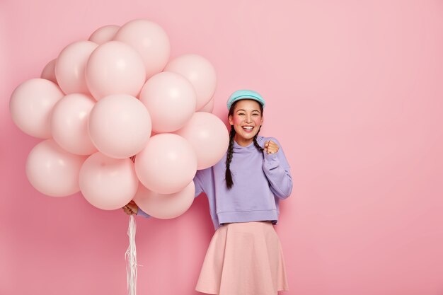 Une fille asiatique heureuse serre le poing de joie, ne peut pas attendre un moment spécial, reçoit les félicitations d'amis pour son anniversaire, porte un gros bouquet de ballons, vêtue de vêtements à la mode