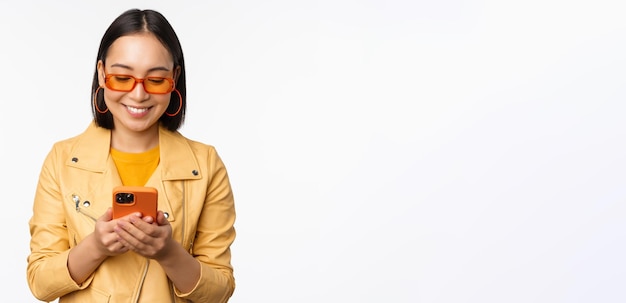 Fille asiatique moderne dans des lunettes de soleil à l'aide de son téléphone portable en souriant et à la pose heureuse contre la Pentecôte