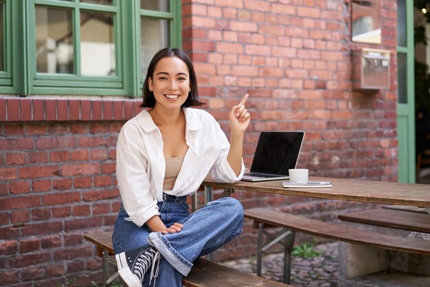 Fille asiatique moderne et élégante avec ordinateur portable assis dans un café à la stupéfaction et pointant vers le coin supérieur droit