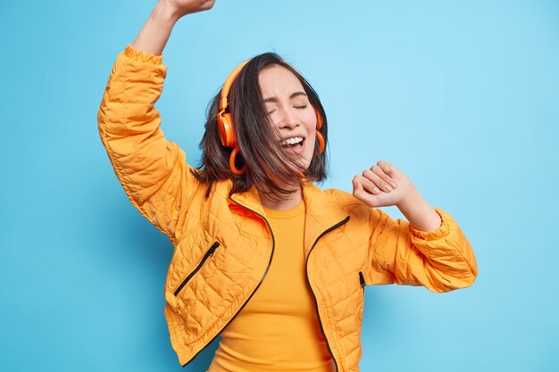 une fille asiatique ravie bouge activement danse insouciante garde les bras levés profite d'une qualité sonore impressionnante via un casque écoute de la musique a les cheveux noirs flottant sur le vent porte une veste orange