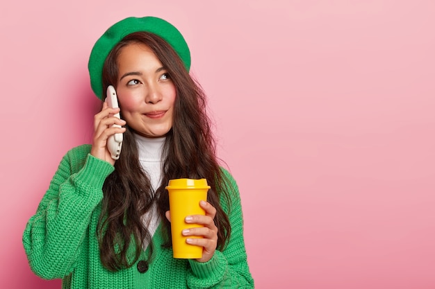 Fille asiatique à la recherche agréable et rêveuse tient le smartphone près de l'oreille, profite d'une conversation agréable tout en buvant du café à emporter, a de longs cheveux noirs, vêtus de vêtements élégants verts