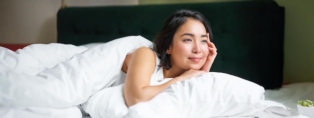 Photo gratuite fille asiatique rêveuse allongée dans son lit le matin recouverte de duver chaud et appuyée sur un oreiller blanc