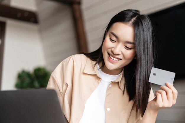 Une fille asiatique souriante achetant en ligne tenant une carte de crédit à la main, faisant du shopping sur un ordinateur portable depuis sa maison vivant...