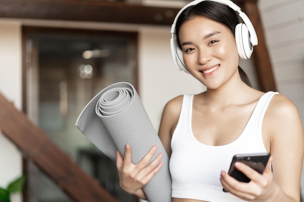 Une fille asiatique souriante dans un casque, écoutant de la musique à la maison et faisant de l'exercice avec un tapis de sol tenant un p...