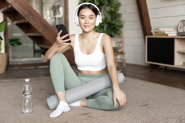 Photo gratuite fille asiatique souriante dans des écouteurs portant des vêtements de sport écoutant de la musique tenant un téléphone portable et un travail...