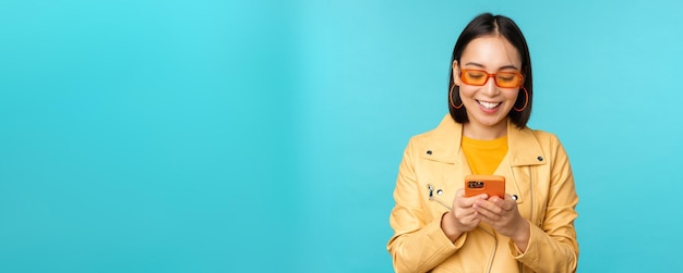 Fille asiatique souriante dans des lunettes de soleil à l'aide d'une application pour smartphone tenant un téléphone portable debout sur une zone bleue