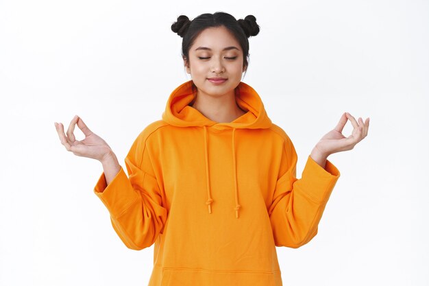 Fille asiatique souriante heureuse et calme, détendue, méditant dans une pose de lotus avec un geste gen, ferme les yeux et sourit paisiblement, se sentant l'esprit tranquille, soulagée après une dure journée d'université