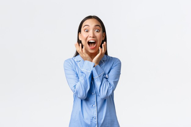 Fille asiatique super heureuse excitée en pyjama bleu se réveillant avec une surprise impressionnante, criant de bonheur en regardant la caméra étonnée, réagit à un cadeau génial, debout sur fond blanc.