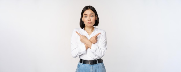 Une fille asiatique triste et confuse ne peut pas décider de pointer les doigts à gauche et à droite en boudant et en haussant les épaules indécise ne peut pas choisir de se tenir debout sur fond blanc