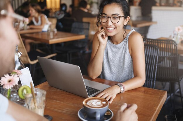 Fille attirée par un gars qui parle en souriant en le regardant amusé admiration rire des histoires drôles Une jolie femme européenne porte des lunettes profite d'une conversation amicale avec un café s'assoit à une table près d'un ordinateur portable