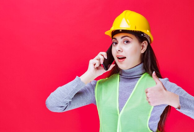 Fille au casque jaune parlant au téléphone et montrant le pouce vers le haut.