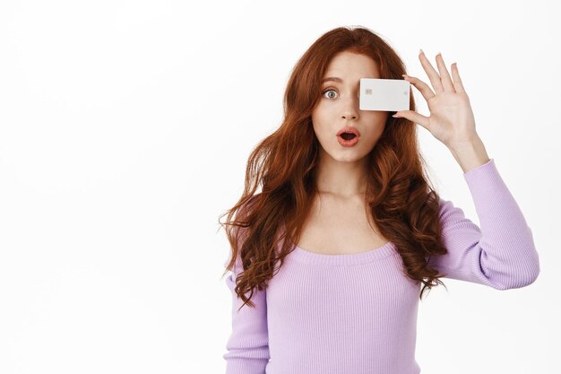 Une fille au gingembre étonnée halète et dit wow, montre une carte de crédit bancaire près de l'œil, promotion d'achats publicitaires, debout en blouse sur fond blanc