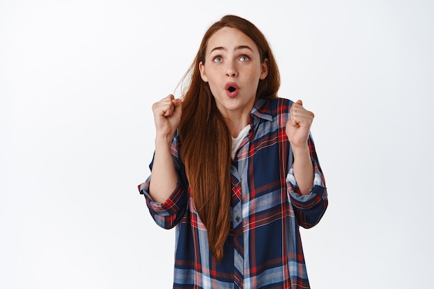 Une fille au gingembre excitée dit wow, dansant d'excitation, regardant l'écran de télévision et chantant, regardant un match de sport et s'enracinant pour l'équipe, debout sur du blanc.