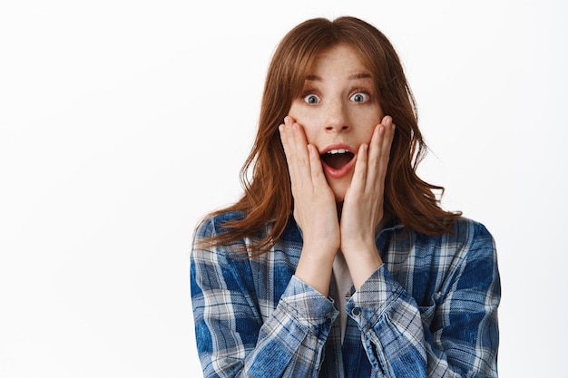 Photo gratuite une fille au gingembre surprise et excitée regarde avec admiration, laisse tomber la mâchoire et haletant étonnée, dit wow, tiens les mains sur les joues, regarde quelque chose d'incroyable et d'étonnant, debout sur fond blanc