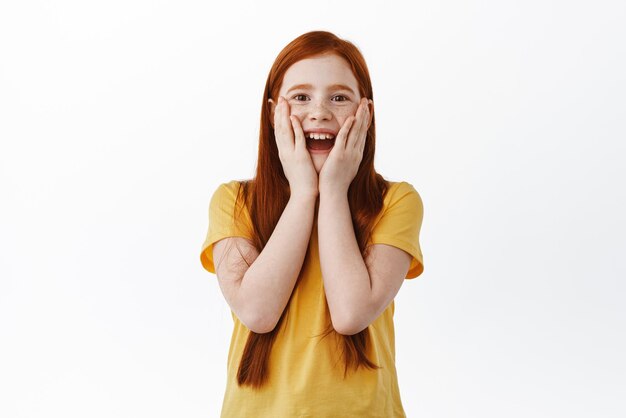 Fille au gingembre surprise avec des taches de rousseur vérifiant quelque chose de génial regardant avec étonnement et joie au cadeau surprise debout heureux sur fond blanc