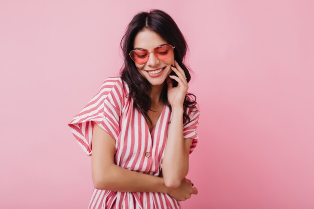 Fille aux cheveux bruns romantique dans des lunettes de coeur à la mode posant avec un sourire timide. Photo intérieure d'une jeune femme gracieuse en tenue d'été et accessoires.