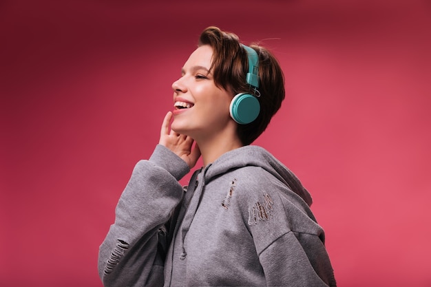 Fille aux cheveux courts de bonne humeur en écoutant la chanson dans les écouteurs. Cheerful woman in hoodie gris sourit et aime la musique sur fond rose isolé