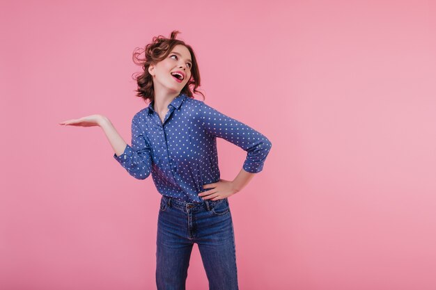 Fille aux cheveux noirs active en chemisier bleu dansant Photo intérieure d'une jeune femme blonde aux cheveux ondulés s'amusant sur un mur rose.