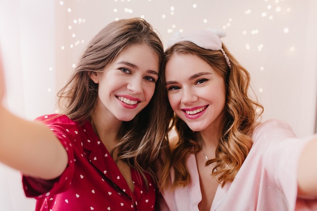 Une fille aux cheveux noirs fascinante porte un costume de nuit rouge faisant un selfie avec une sœur souriante. Photo intérieure de deux jolies dames en pyjama mignon se prenant en photo.