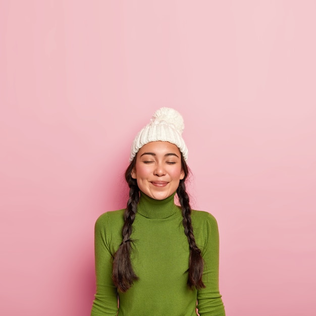 Une fille aux cheveux noirs heureuse garde les yeux fermés, pense à une rencontre agréable avec un ami, porte un chapeau blanc et un col roulé vert, se tient contre un mur rose