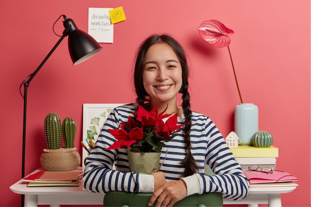 Fille aux cheveux noirs positive tient une fleur en pot rouge, pose contre un espace de coworking confortable