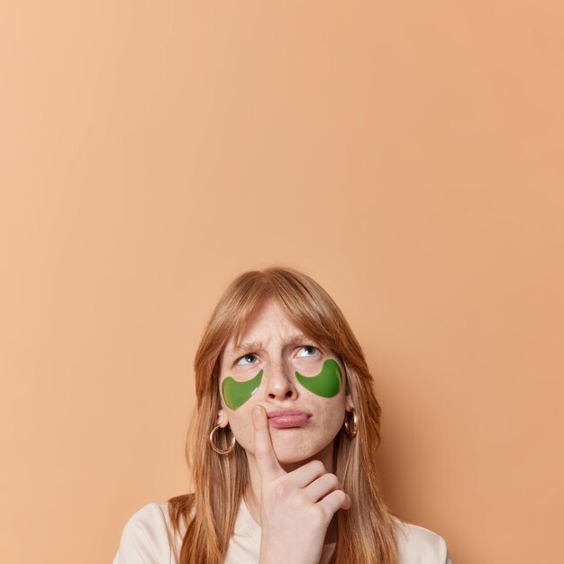 Une fille aux cheveux rouges mécontente fronce les sourcils et regarde au-dessus garde le doigt près des lèvres pense à quelque chose applique des patchs de beauté pour les soins de la peau isolés sur fond marron copie vierge espace au-dessus