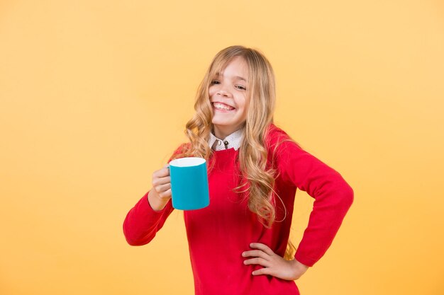Fille aux longs cheveux blonds en pull rouge tenir la tasse. sourire d'enfant avec une tasse bleue sur fond orange. soif, concept de déshydratation. santé et boisson saine. pause thé ou café.