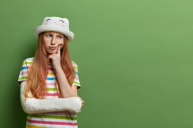 Fille aux taches de rousseur pensif aux cheveux roux, garde le doigt sur la joue, a déplu à l'expression, a le bras cassé, ne peut pas jouer avec les enfants en plein air, isolé sur un mur vert, espace vide pour la promo