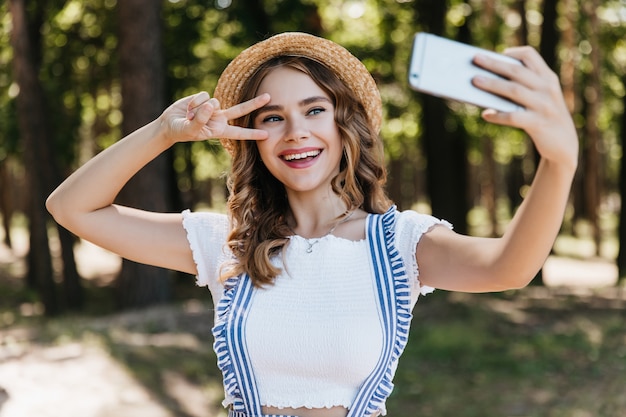 Fille aux yeux bleus extatique au chapeau à l'aide de téléphone pour selfie. Dame de bonne humeur aux cheveux bouclés posant avec signe de paix dans la forêt.