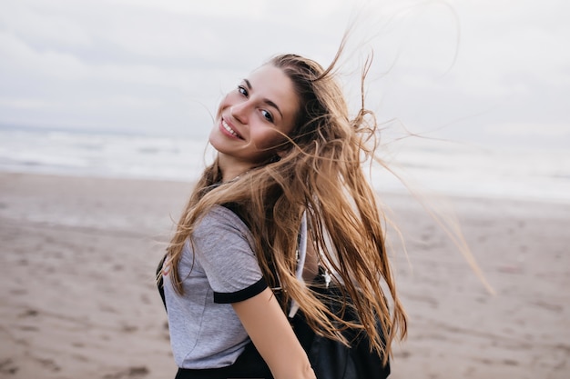 Fille aux yeux noirs fascinante s'amusant à la plage le matin du week-end et posant. Modèle féminin aux cheveux bruns excité profitant de l'extérieur